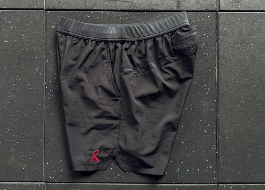 Side view of black training shorts with a red embroidery on the left leg, a small 'R' logo on the right leg, and a rear mesh panel, displayed against a dark textured background.