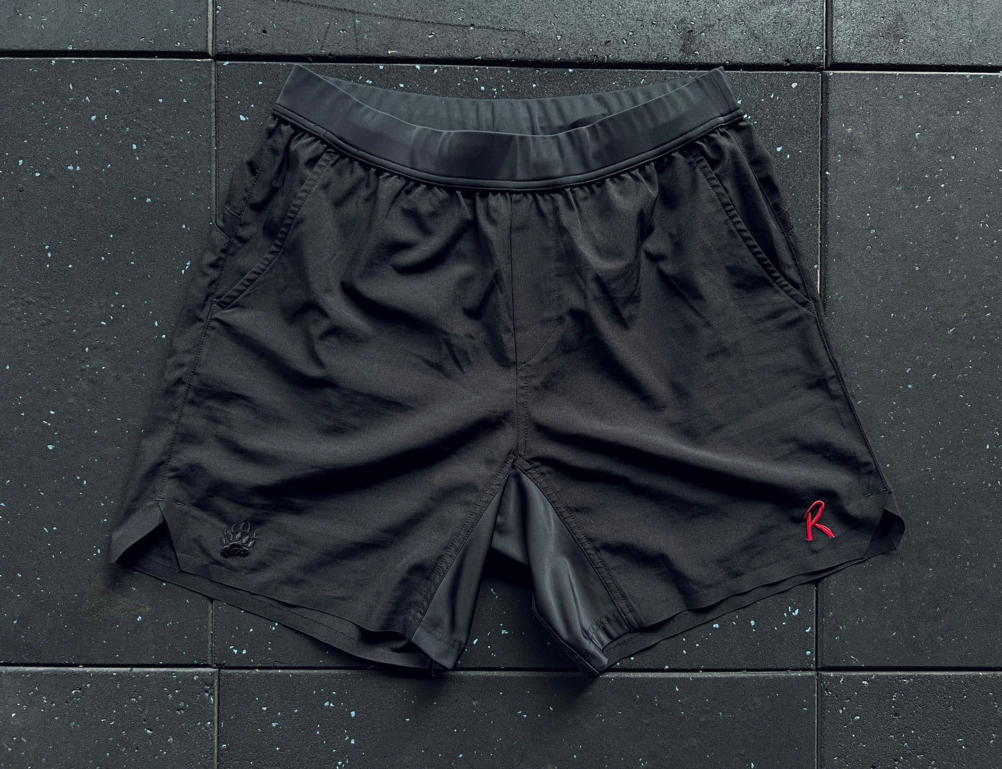 Black training shorts with a red embroidery on the left leg, a small 'R' logo on the right leg, and a rear mesh panel, displayed against a dark textured background.
