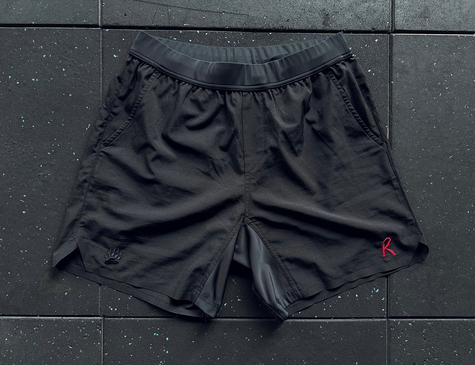 Black training shorts with a red embroidery on the left leg, a small 'R' logo on the right leg, and a rear mesh panel, displayed against a dark textured background.