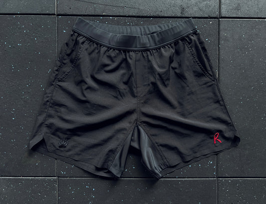 Black training shorts with a red embroidery on the left leg, a small 'R' logo on the right leg, and a rear mesh panel, displayed against a dark textured background.
