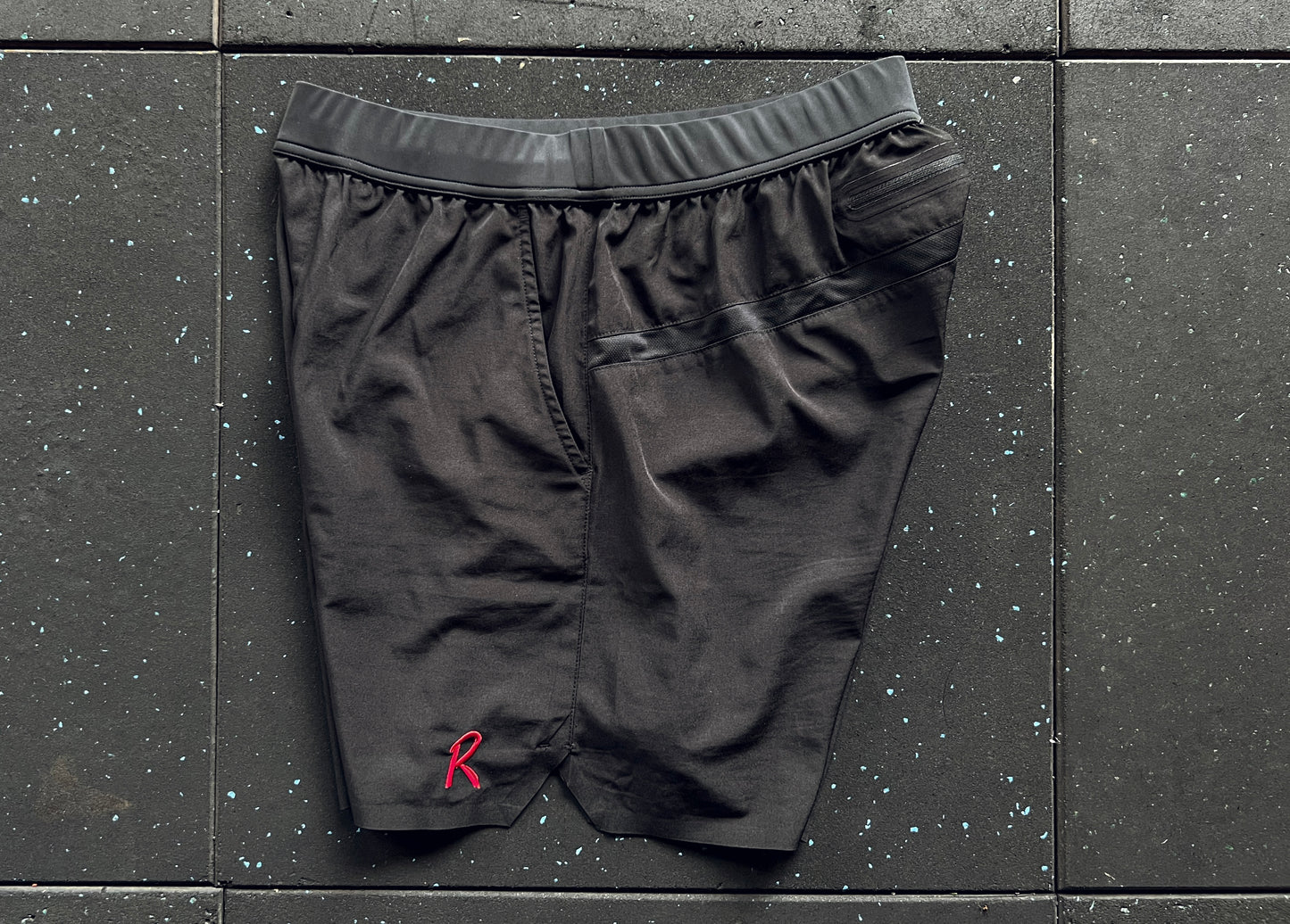 Side view of black training shorts with a red embroidery on the left leg, a small 'R' logo on the right leg, and a rear mesh panel, displayed against a dark textured background.